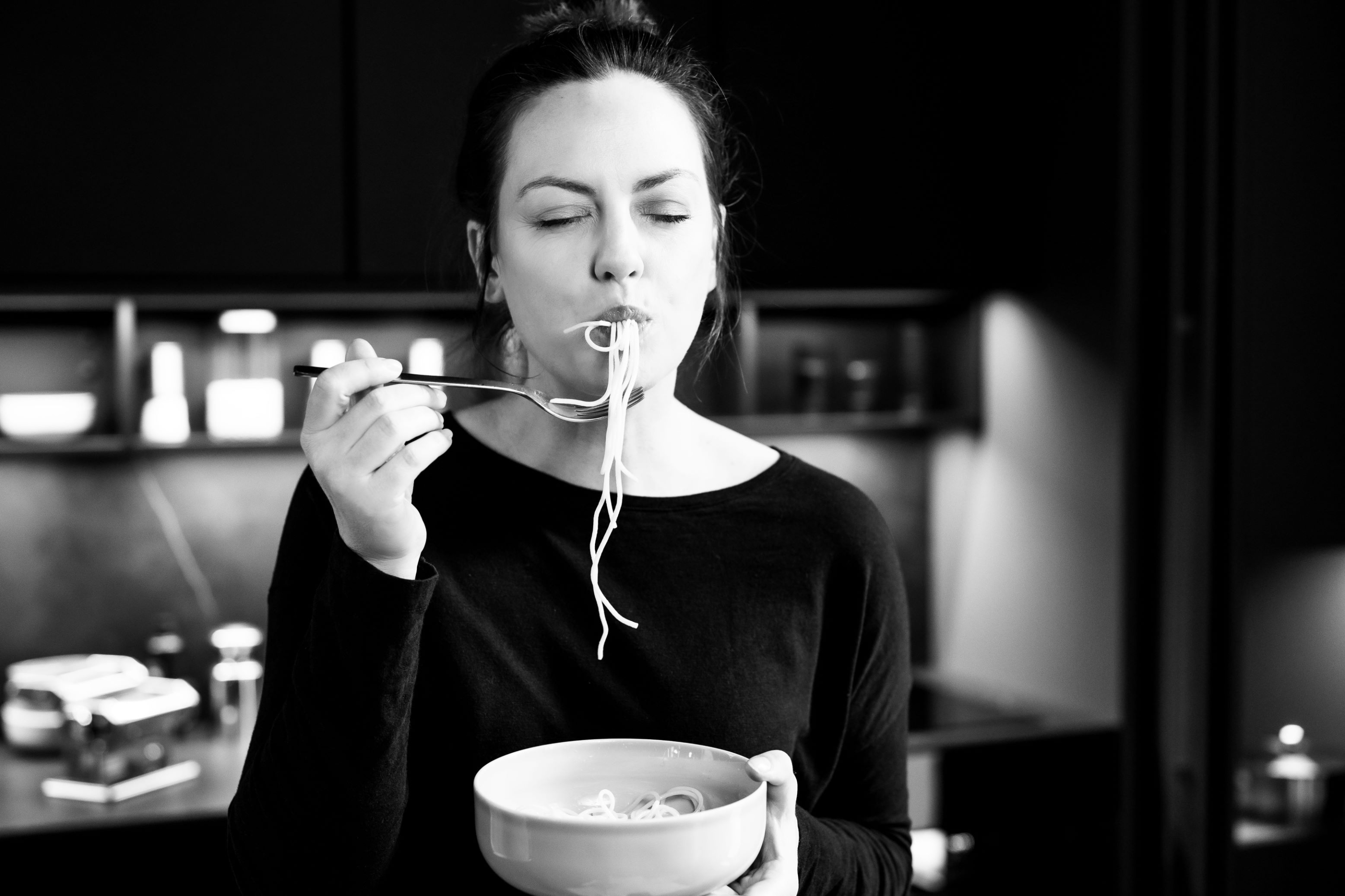 Werbemotiv der Linie dk. Bild in sw, Frau hält Schüssel und isst daraus Spaghetti, ihre Augen sind geschlossen.