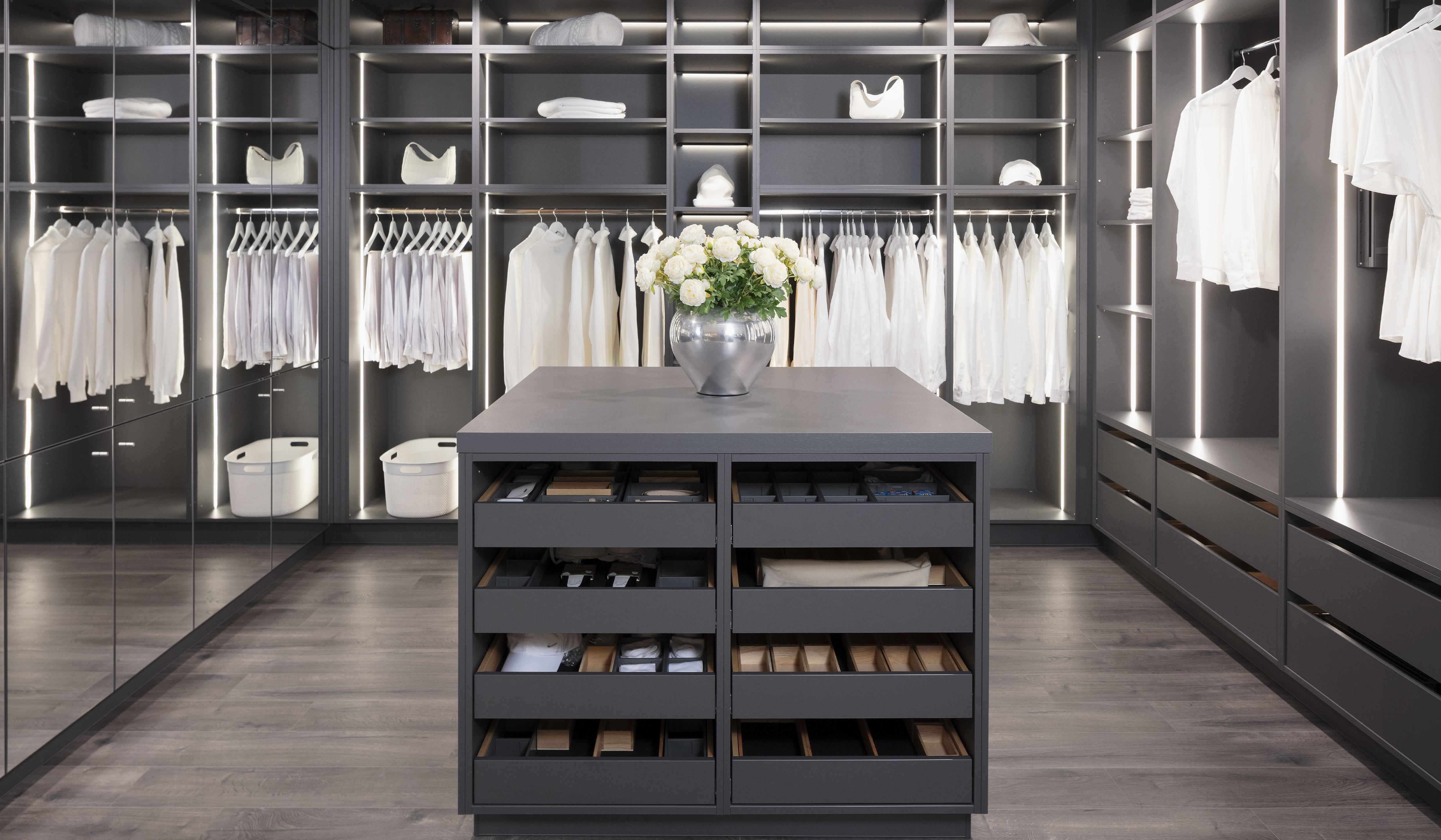 ein komplettes ANkleidezimmer mit Spiegelwand, Schrankwand gefüllt mit Kleidung sowie Insel mit Schubladen für Schuhe und Gürtel. 